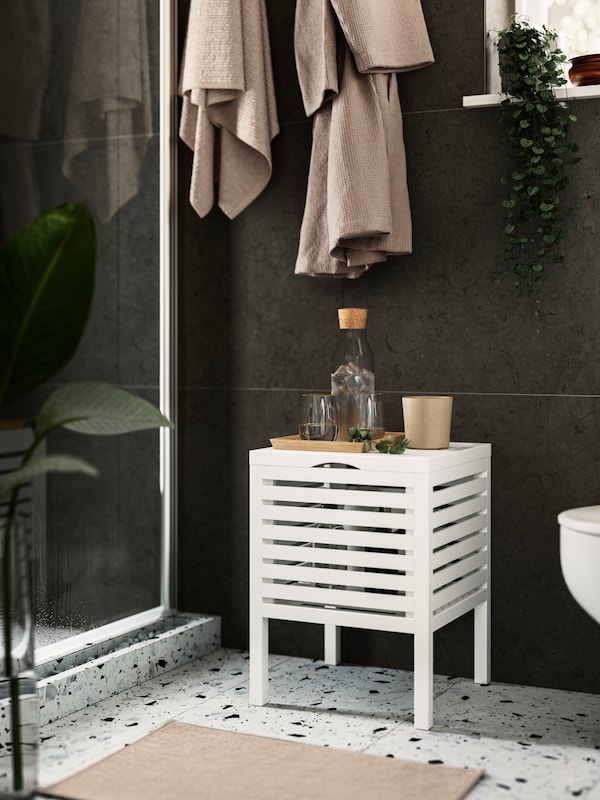 A MUSKAN storage stool in white with glasses of water on top. A green plant and two beige bathrobes on the wall behind it.
