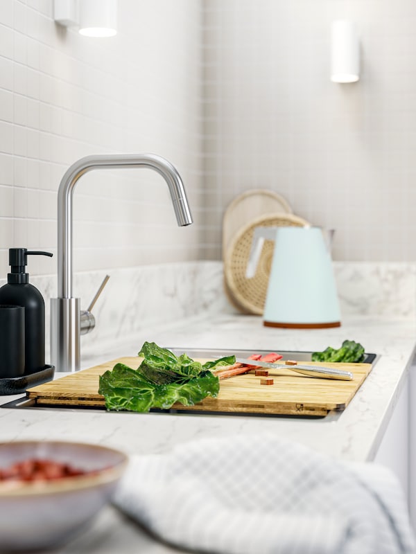 A kitchen countertop with a NORRSJÖN chopping board, fresh vegetables, and a kettle, all set against a tiled backsplash.