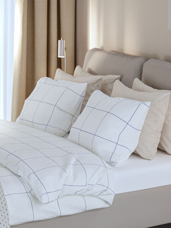 A hotel room with a SAGESUND upholstered bed made with BREDVECKLARE bedding next to a bedside table in oak veneer.