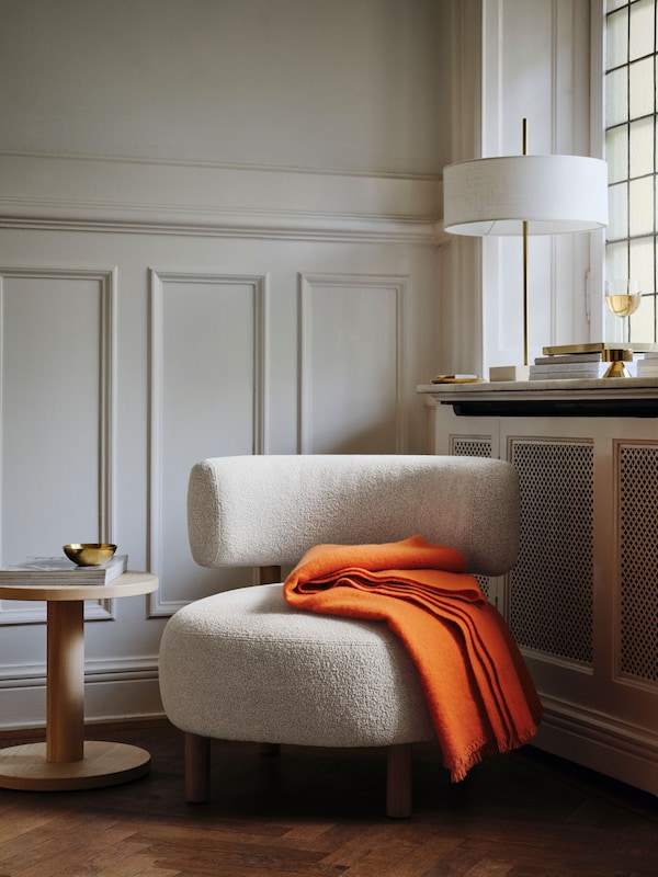 A beige STOCKHOLM 2025 armchair by a window in the corner of a room. A bright orange throw rests on the armchair.