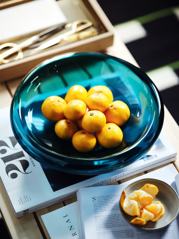 A dark blue-turquoise STOCKHOLM 2025 bowl with fruit next to an oak veneer tray and a grey serving plate with peeled fruit.